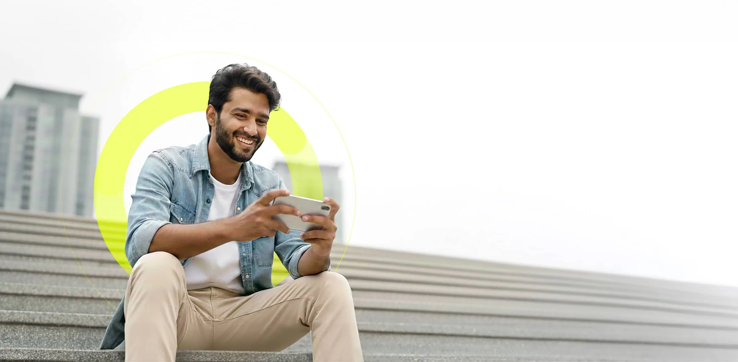 Man on steps with beard wearing an open denim shirt and white t-shirt is sat outside on some steps looking at his mobile phone.