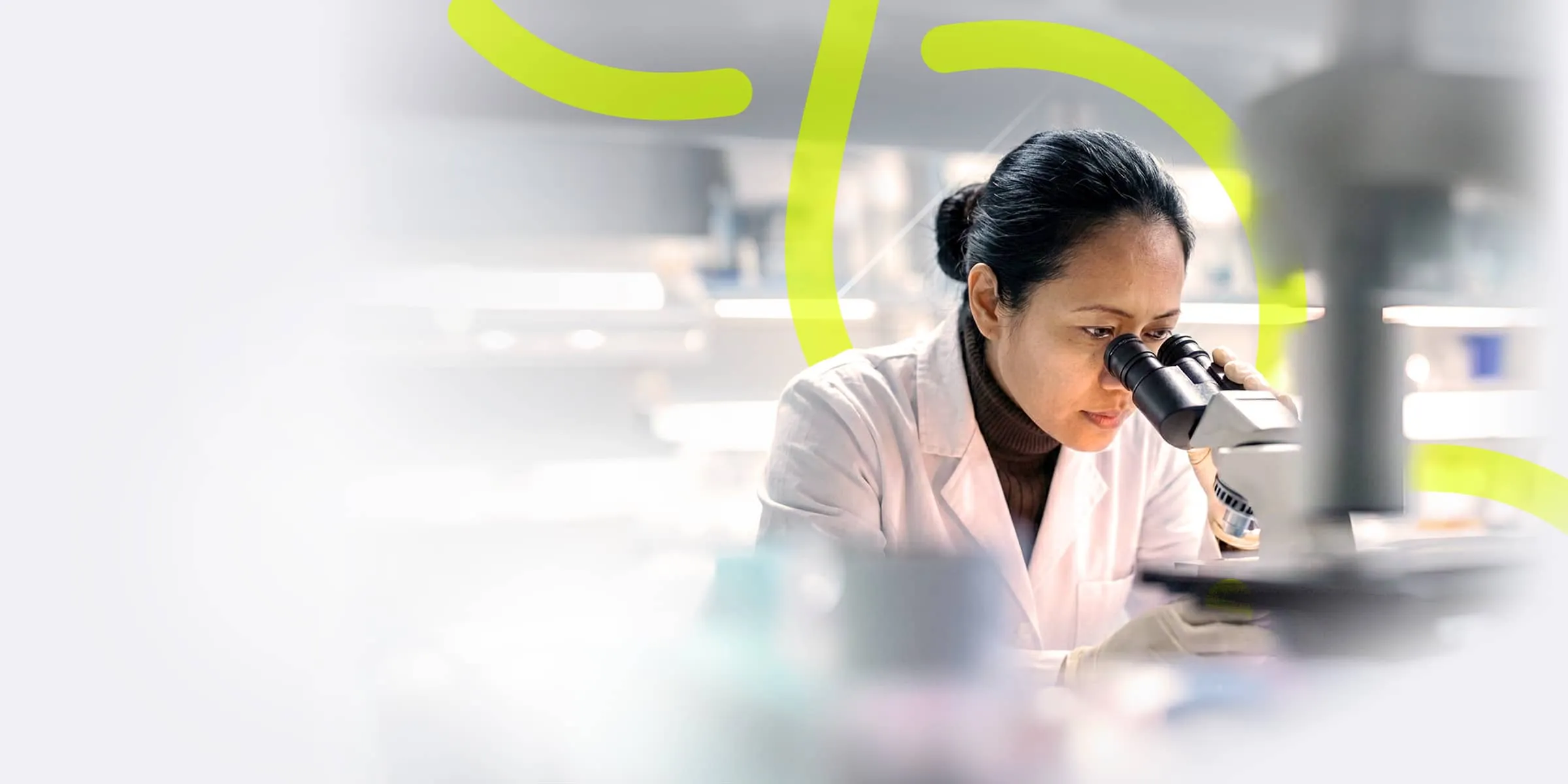 woman wearing a white lab coat is looking into a powerful microscope
